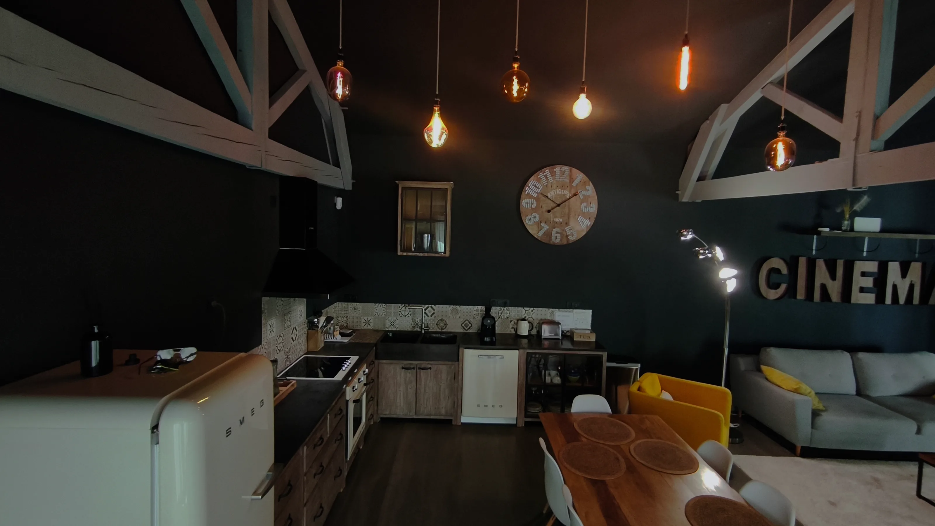 La cuisine sous la charpente peinte, ampoules suspendues et horloge de bois